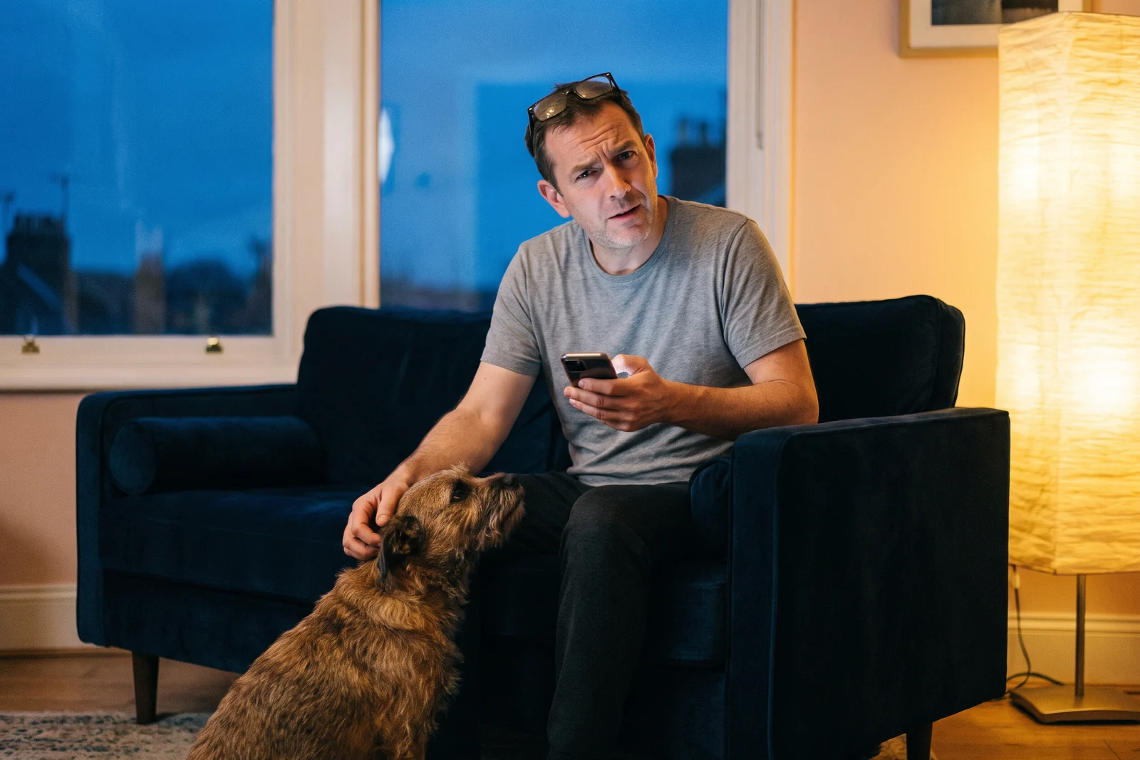 In a living room in the early evening, a man in a gray T-shirt with his glasses pushed up on his forehead scratches a waking dog behind the ear with one hand and holds his phone in the other, its screen lighting his face as he waits for an answer.
