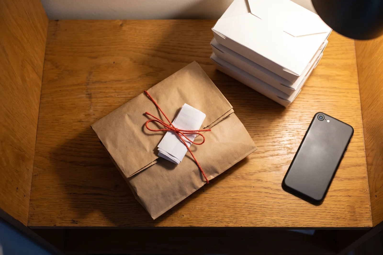 Top-down on the pickup shelf: a paper bag folded shut and tied with twine, a folded ticket tucked under the twine, a stack of unlabeled white containers beside it and a phone lying face down in the warm lamplight.