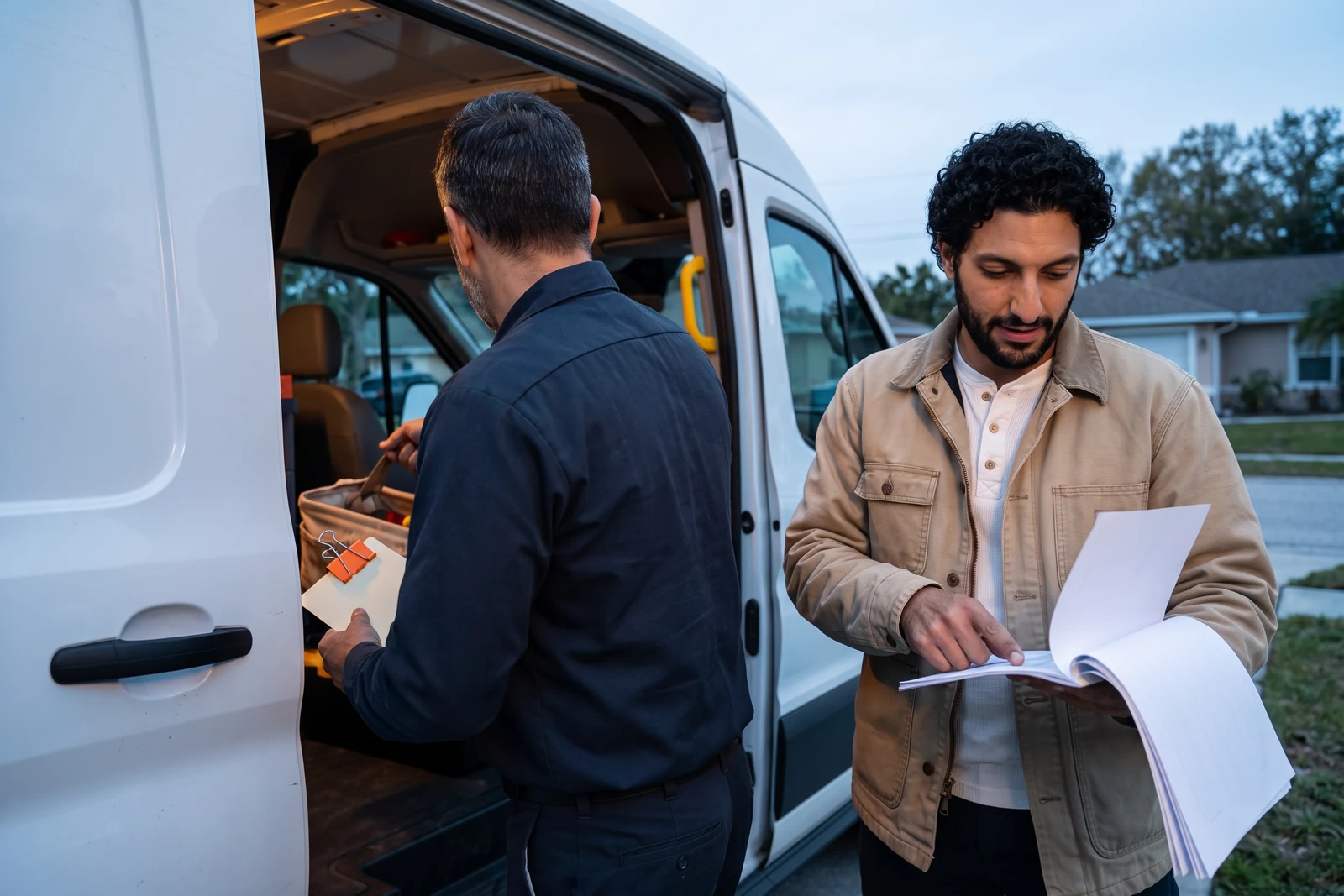 At first light in a driveway, the owner swings a toolbox into the open side door of his work van with one hand and takes the orange clip off a blank card with the other, while Avery, in a sand-colored work jacket, walks beside him holding open a stapled set of blank pages and pointing to a line.