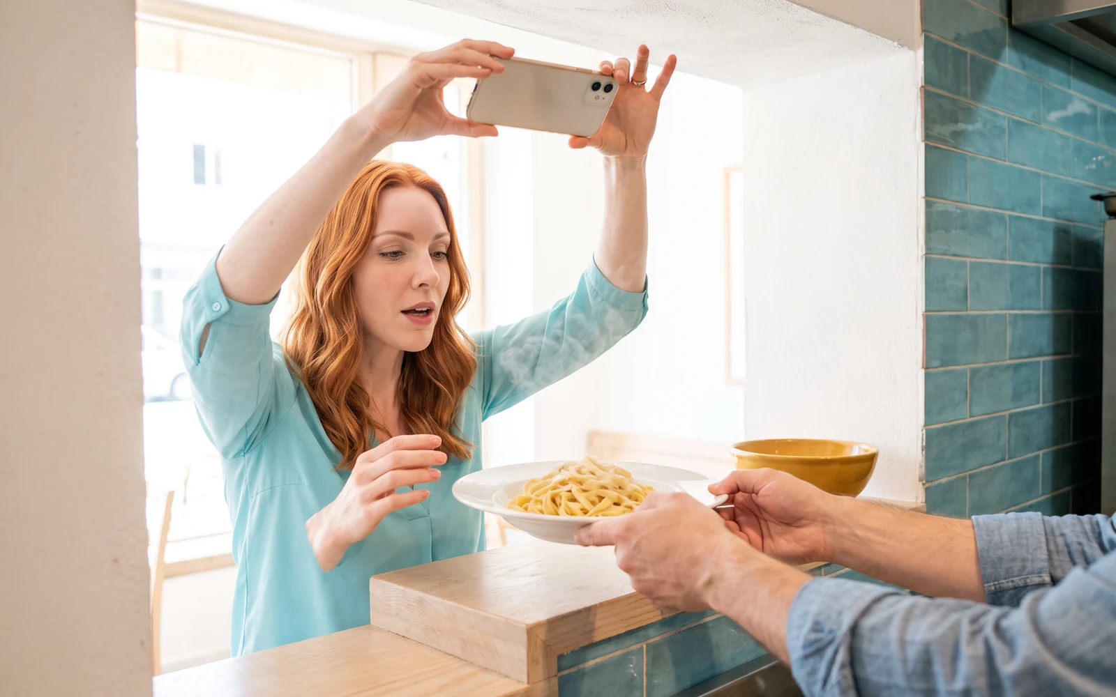 Afternoon at the kitchen pass: Sloane, the AI SMM manager, lifts her phone high to photograph a steaming plate of fettuccine that a cook’s hands hold out through the pass.