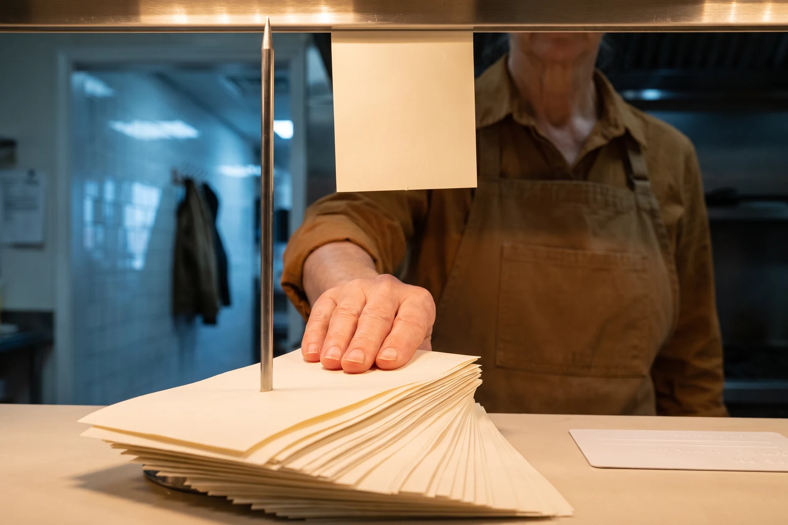 Seen from counter height, a steel spike pierces the frame from below: a hand in a work apron presses one more blank ivory slip down onto the fan already impaled there, a waiting coat blurred beyond the doorway.