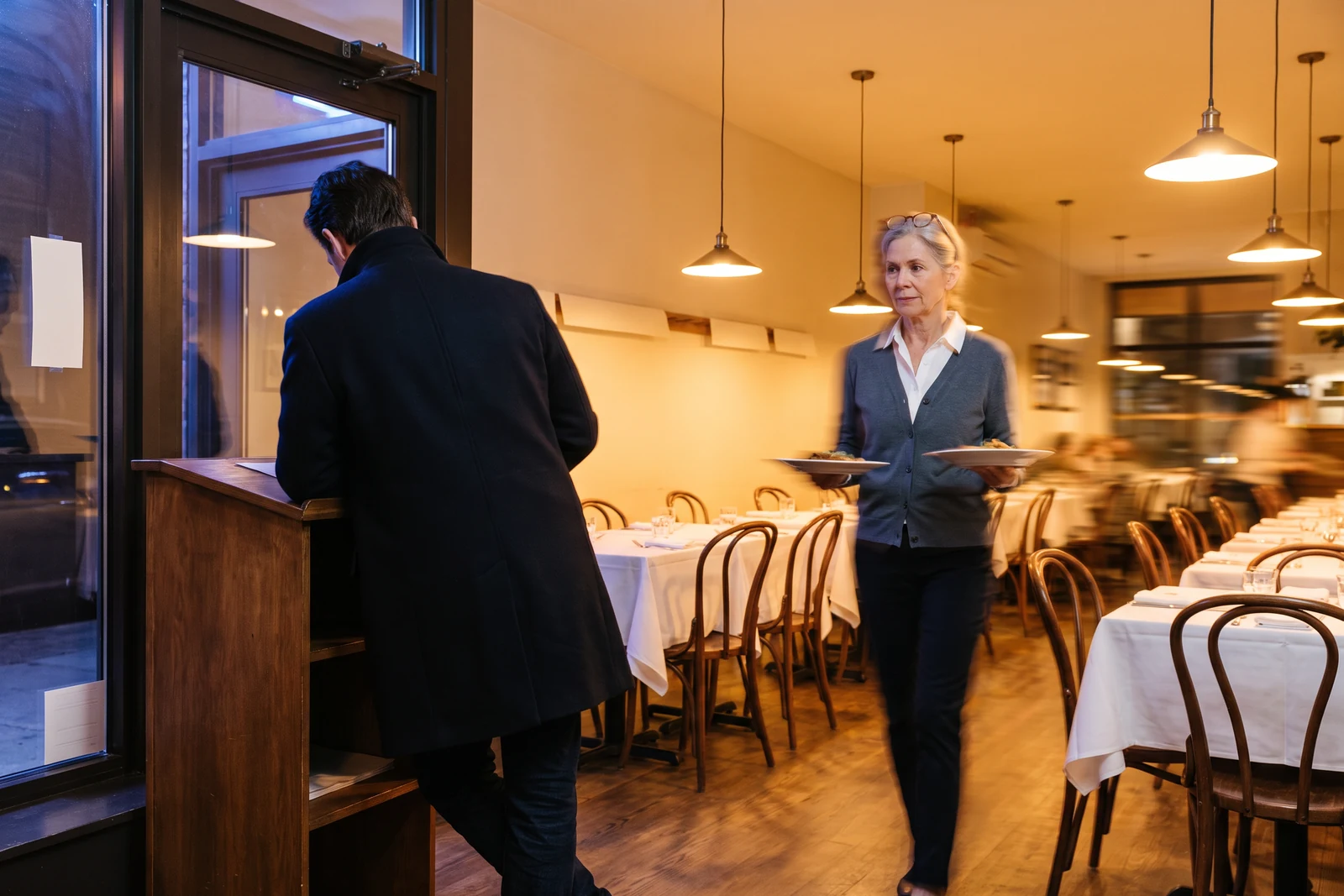 At the front door a guest in a coat leans on the hostess stand with both hands, sharp against the warm dining room blurred behind him; there is nobody on the other side of the stand, and the owner is far away in the room with two plates.