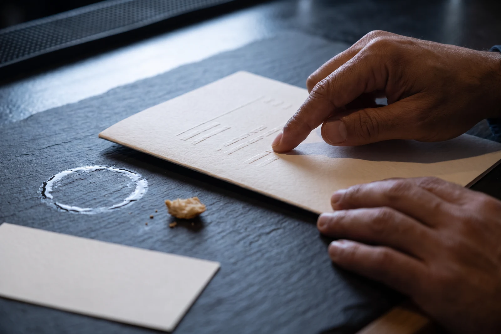 Close along the slate counter: a fingertip slides along one line of a blind-embossed menu card beside a wet ring from a glass, with an untouched blank slip at the edge of the frame.