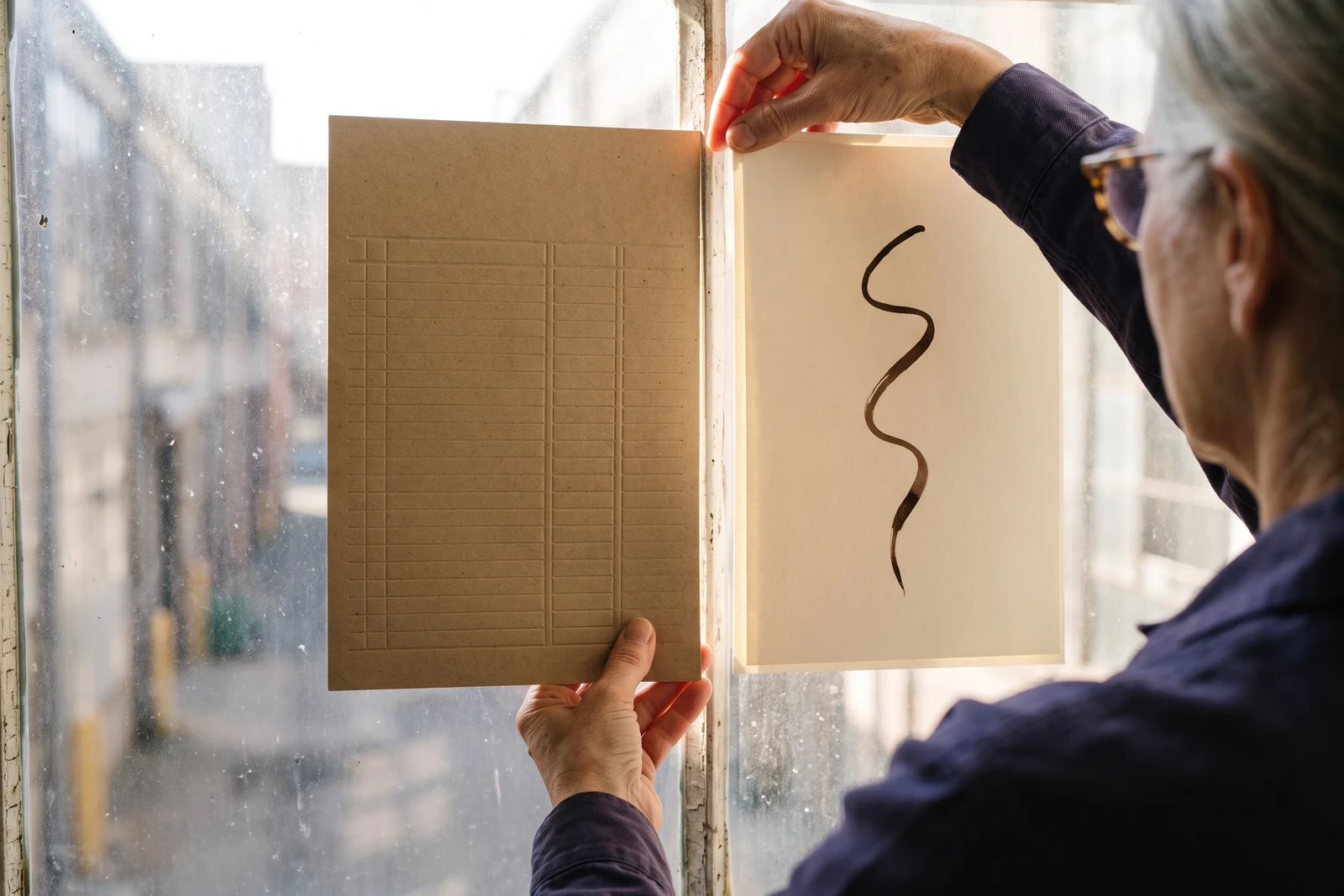 Margaret holds a kraft delivery record and a cream card side by side against a tall window to compare them in the light.