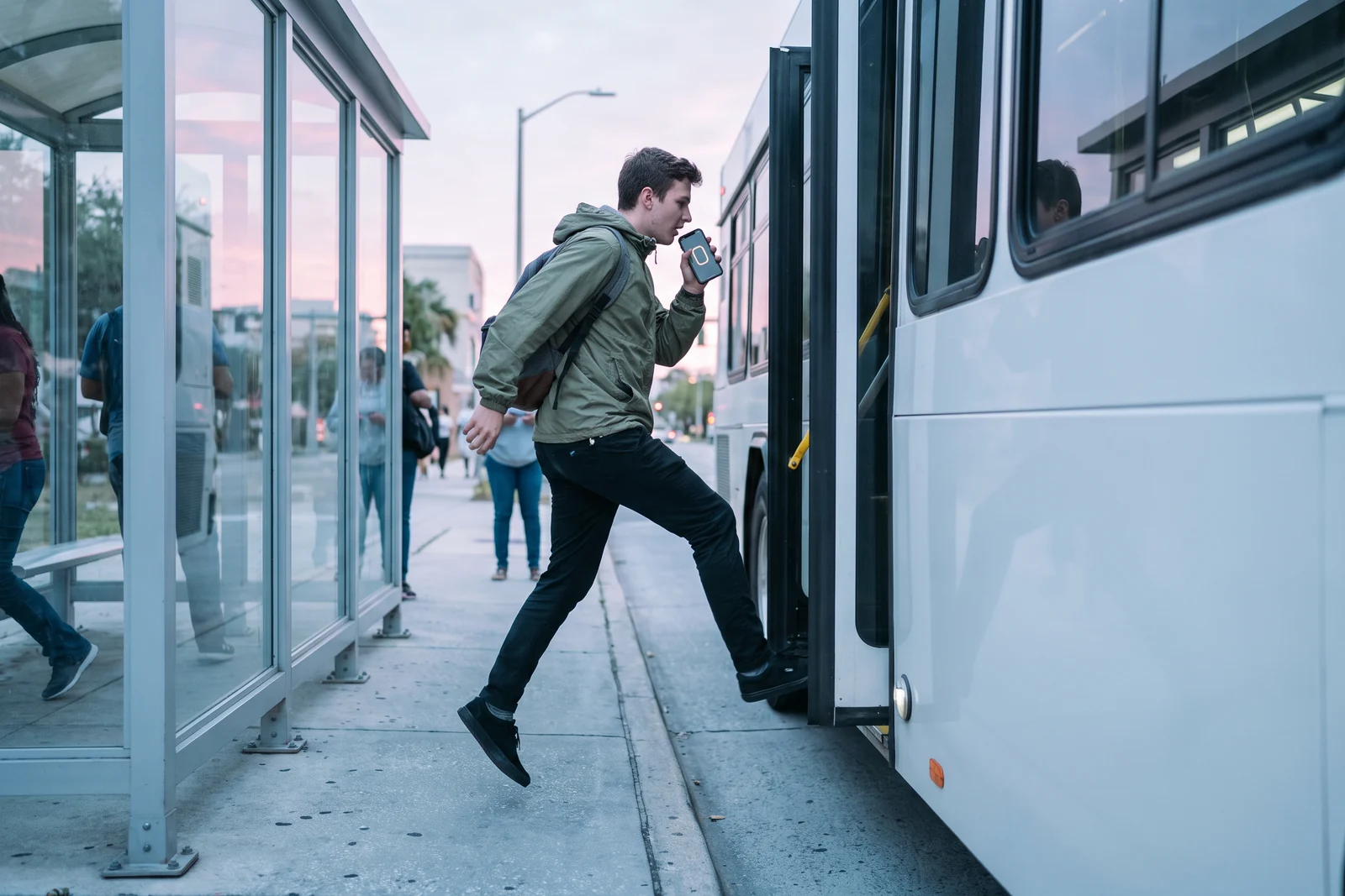 Early morning at a Tampa bus stop: a young man in an olive windbreaker jumps onto the bus step, one hand on the rail, dictating a question into his phone as the door closes.