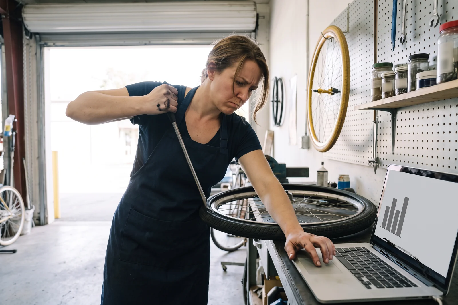 In a bike repair shop, the owner in a navy apron leans her weight on a tire lever and scrolls a laptop of unlabeled bars with a greasy knuckle.