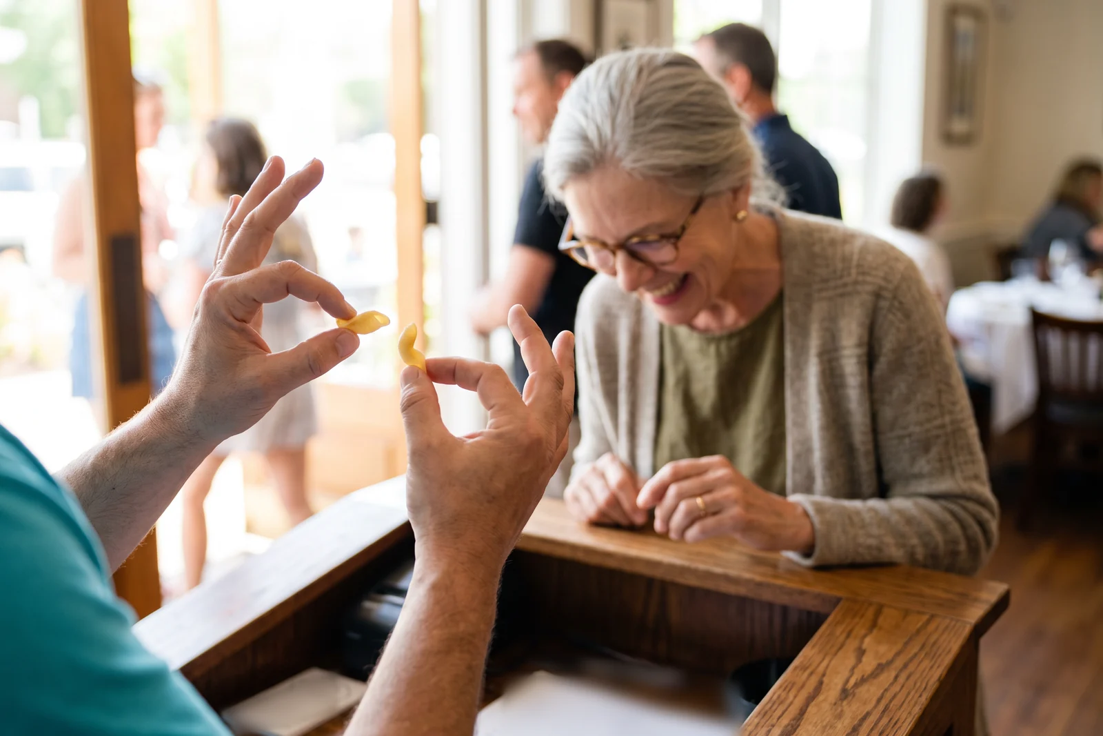 At the host stand at lunch, a guest shapes a small pasta with “little ears” in the air with both hands while Margaret leans in, laughing as she recognizes orecchiette.
