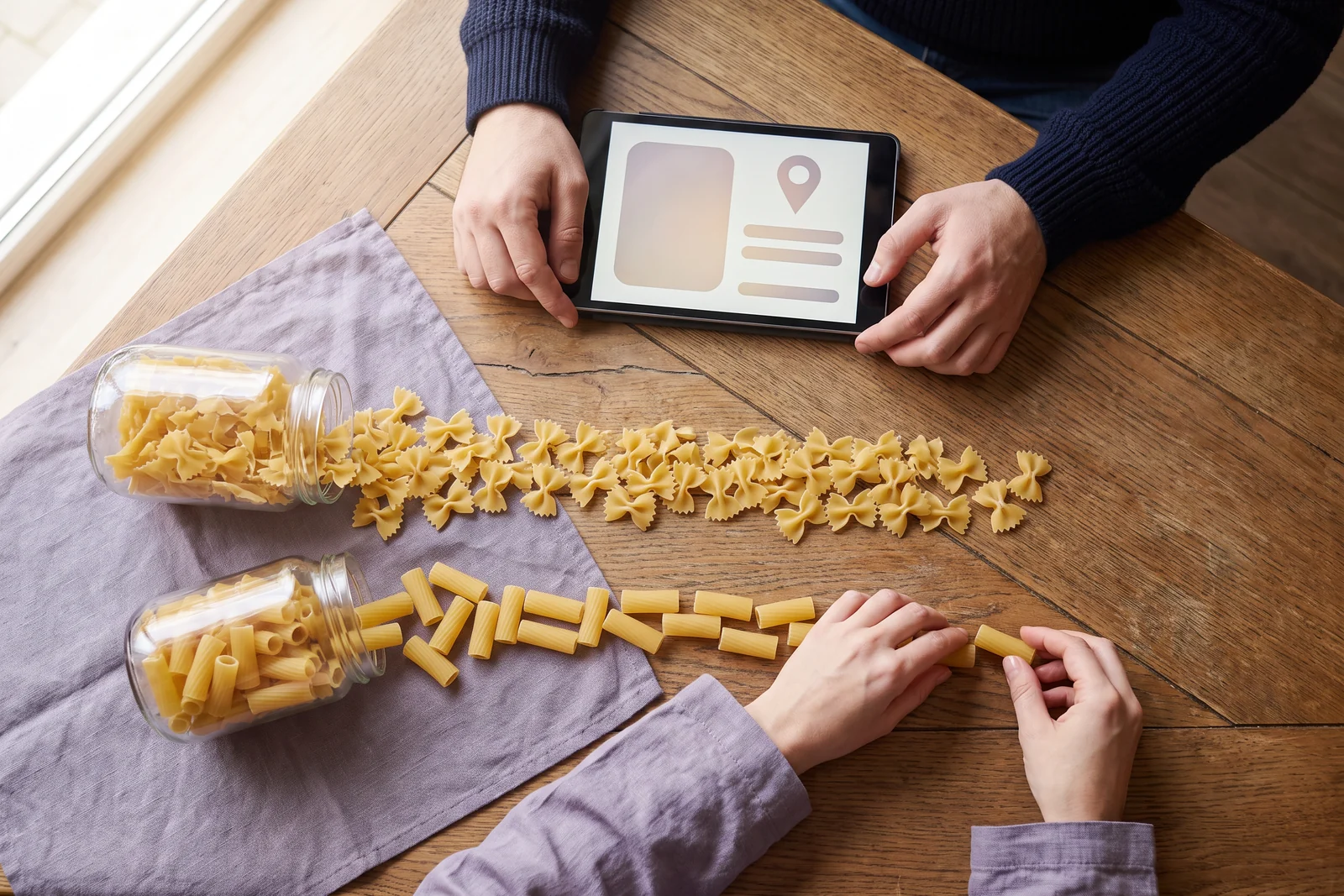 Top-down on a wooden table a month later: two jars tipped out into lines of pasta, the rigatoni line now longer but still shorter than the farfalle; Miles, the AI Business Advisor, turns a tablet toward Margaret as she adds the last rigatoni.
