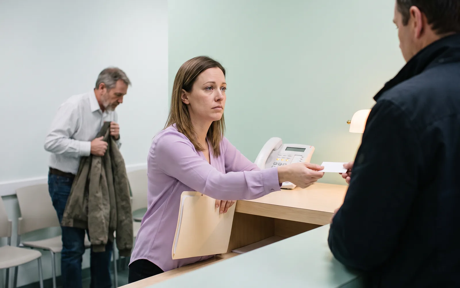 Late morning at a dental front desk: the receptionist reaches across the counter to hand a card back while three phone lines glow on the desk phone beside her, and a patient in the waiting area stands up to leave.